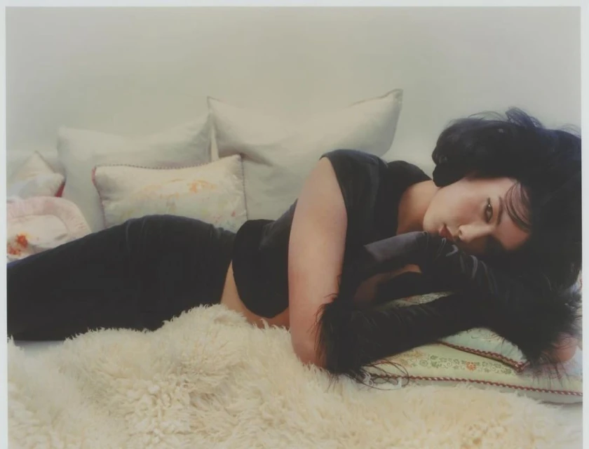 Woman with dark hair lies on a white fluffy rug, with decorative pillows behind her. She wears a black top with furry black arm coverings.