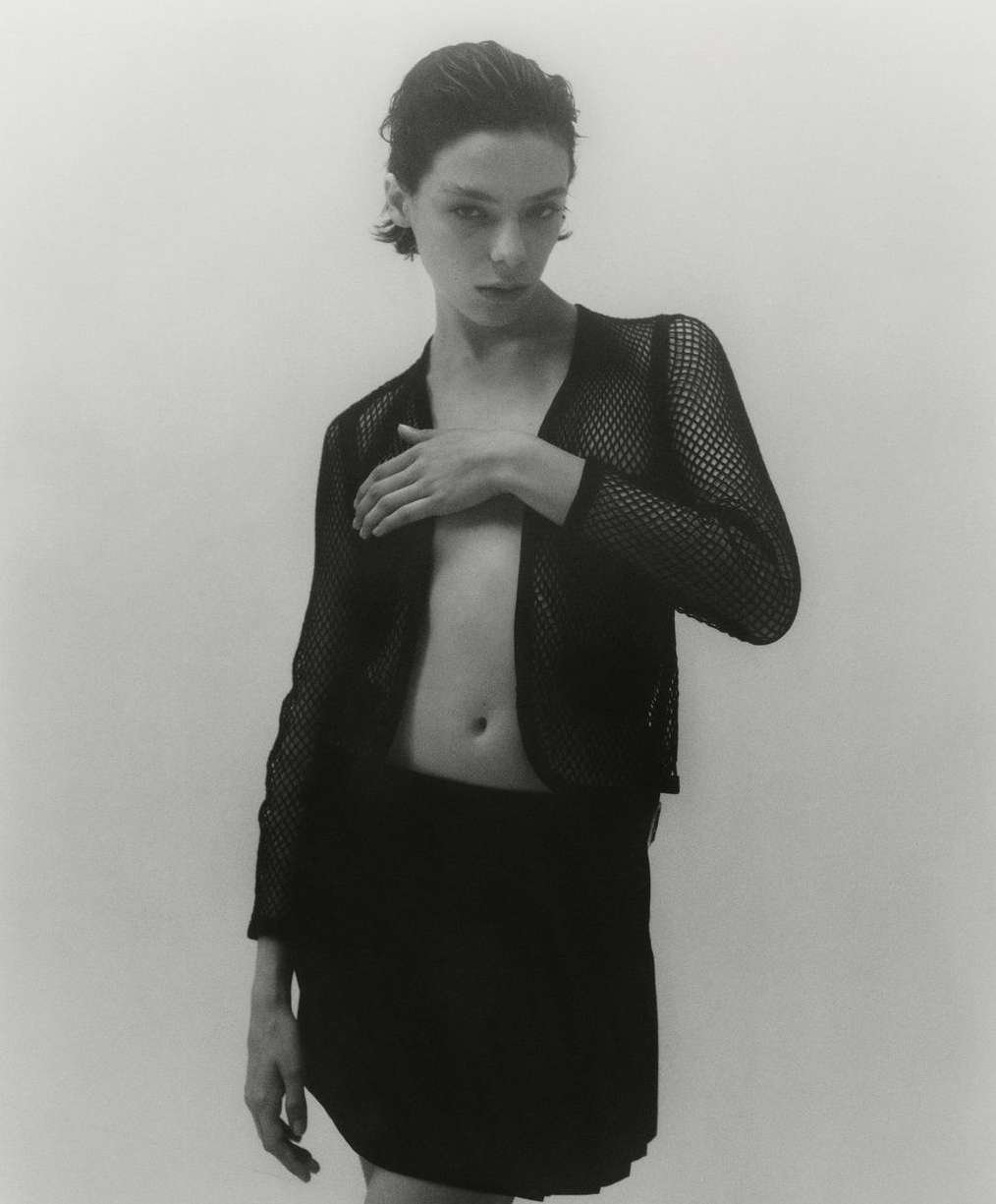 Monochrome portrait of a woman in a mesh top and skirt, posing indoors against a white backdrop, emphasizing her minimalist style and composition.