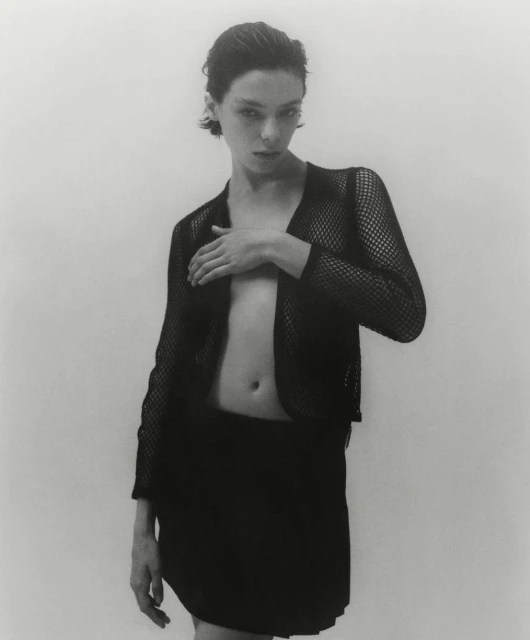 Monochrome portrait of a woman in a mesh top and skirt, posing indoors against a white backdrop, emphasizing her minimalist style and composition.