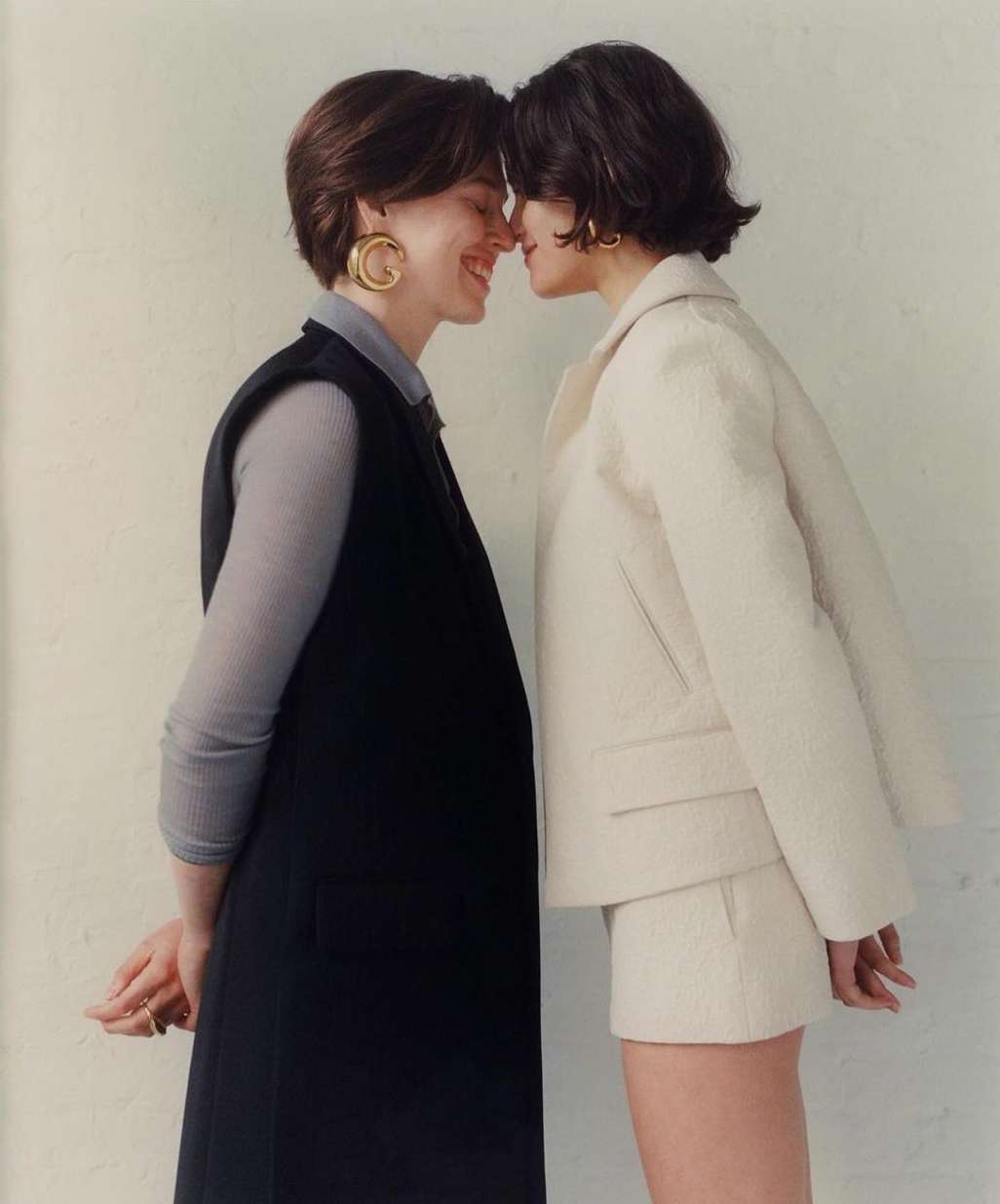 Two short-haired women standing face to face with eyes closed, one in a black vest and one in a white jacket