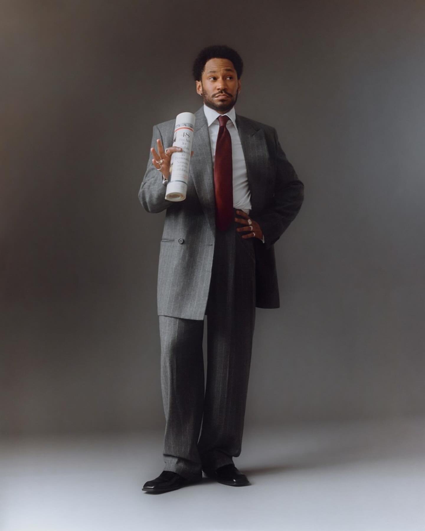 Full-length portrait of an adult male in a gray suit, red tie, and black shoes, holding rolled paper against a gray background.