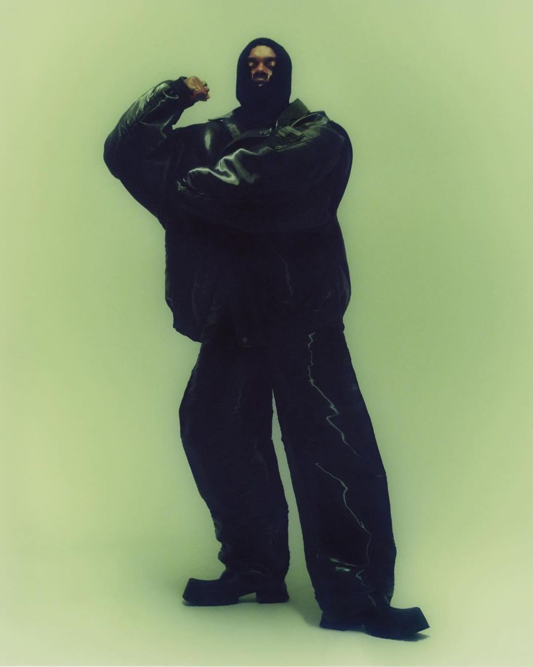 Man wearing a dark oversized leather jacket and matching trousers with head covering against a pale green background