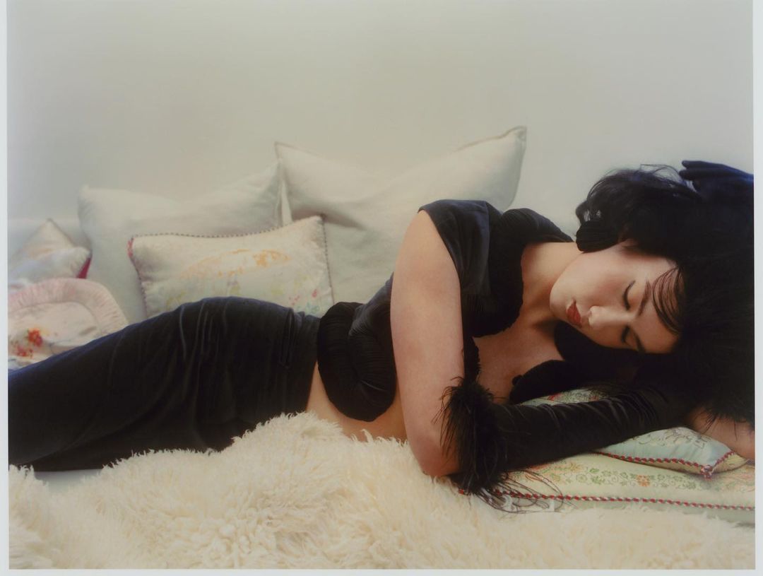 Woman with dark hair wearing a dark top and pants, laying on a white faux fur rug with decorative pillows, against a white wall.