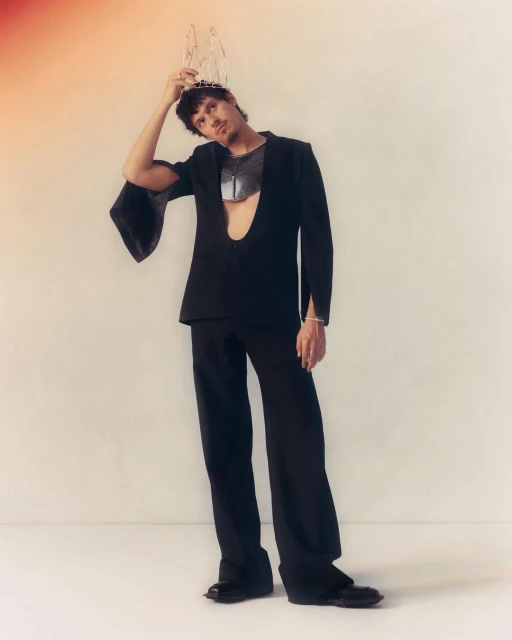 A young adult man wearing a black suit and decorative crown poses in a studio against an off-white background. Fashion shot, portrait orientation.