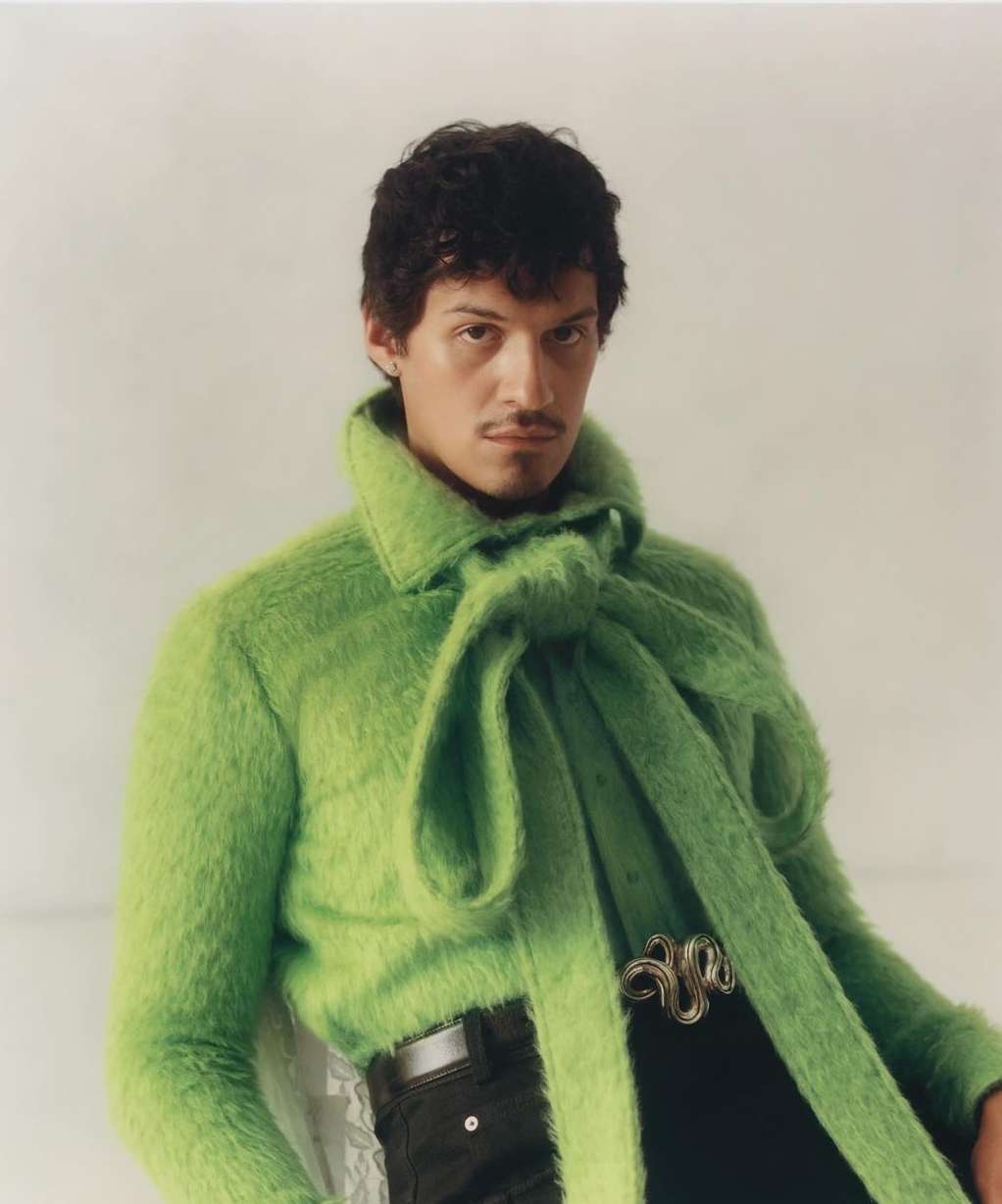 Portrait of a dark-haired man wearing a bright green fuzzy sweater with a tied scarf collar and dark pants, posing against a white background.