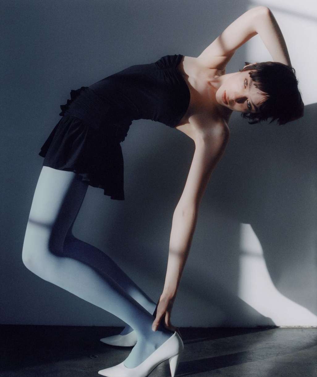 A woman in a black dress and light blue stockings arches her back in a studio setting, creating a striking visual composition.