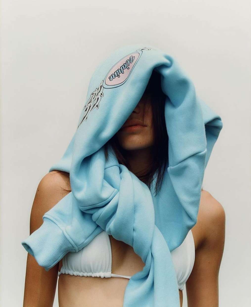 Adult female with light skin wearing a bikini top and a light blue hoodie draped over her head against an off-white background.