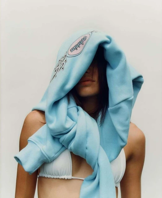 Adult female with light skin wearing a bikini top and a light blue hoodie draped over her head against an off-white background.