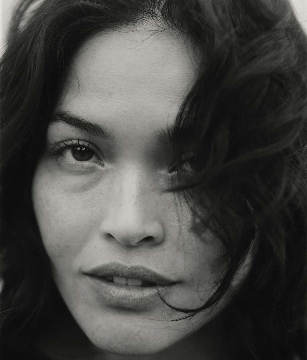 Close-up portrait of an adult female with dark hair, brown eyes, and fair skin. The shot is in black and white with soft lighting.