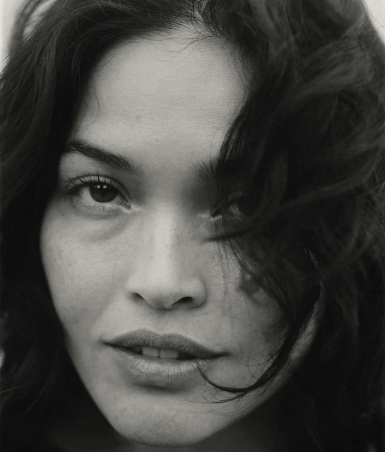 Close-up portrait of an adult female with dark hair, brown eyes, and fair skin. The shot is in black and white with soft lighting.