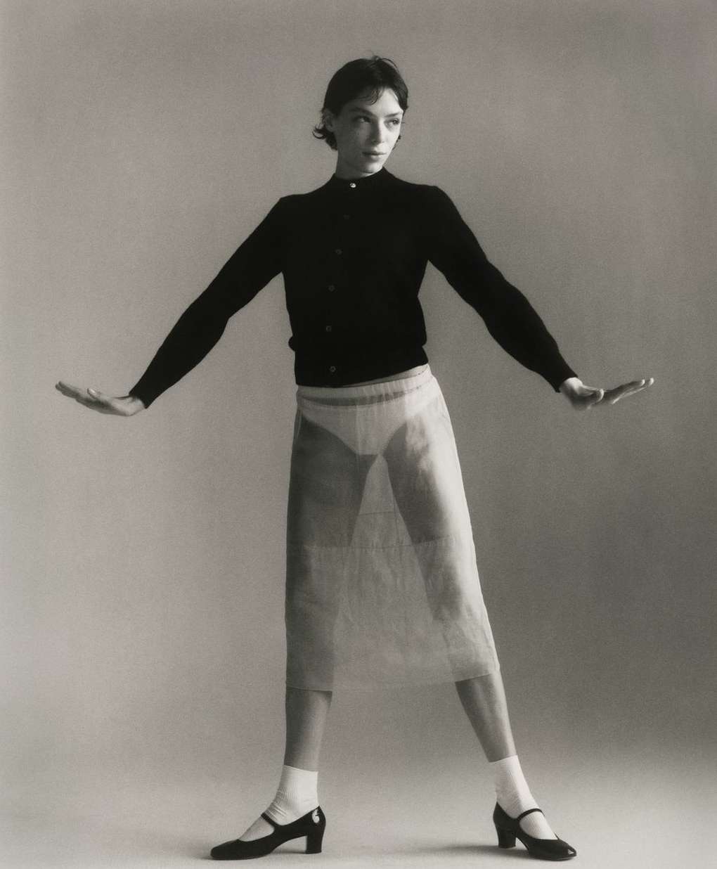 A slender adult woman stands in a photo studio wearing a black cardigan, sheer skirt, white socks, and dark Mary Jane shoes.