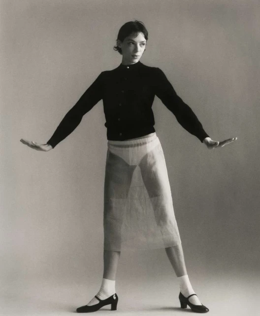 A slender adult woman stands in a photo studio wearing a black cardigan, sheer skirt, white socks, and dark Mary Jane shoes.