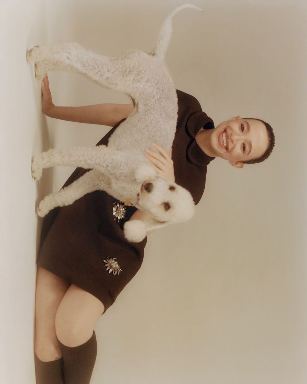 Smiling woman in a brown dress holding a white poodle in a studio setting against a white background for a fashion editorial.