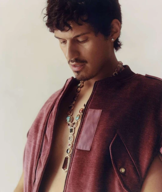 Close-up of a young adult male wearing a maroon sleeveless jacket with a large jeweled necklace, posing in a brightly lit indoor space.