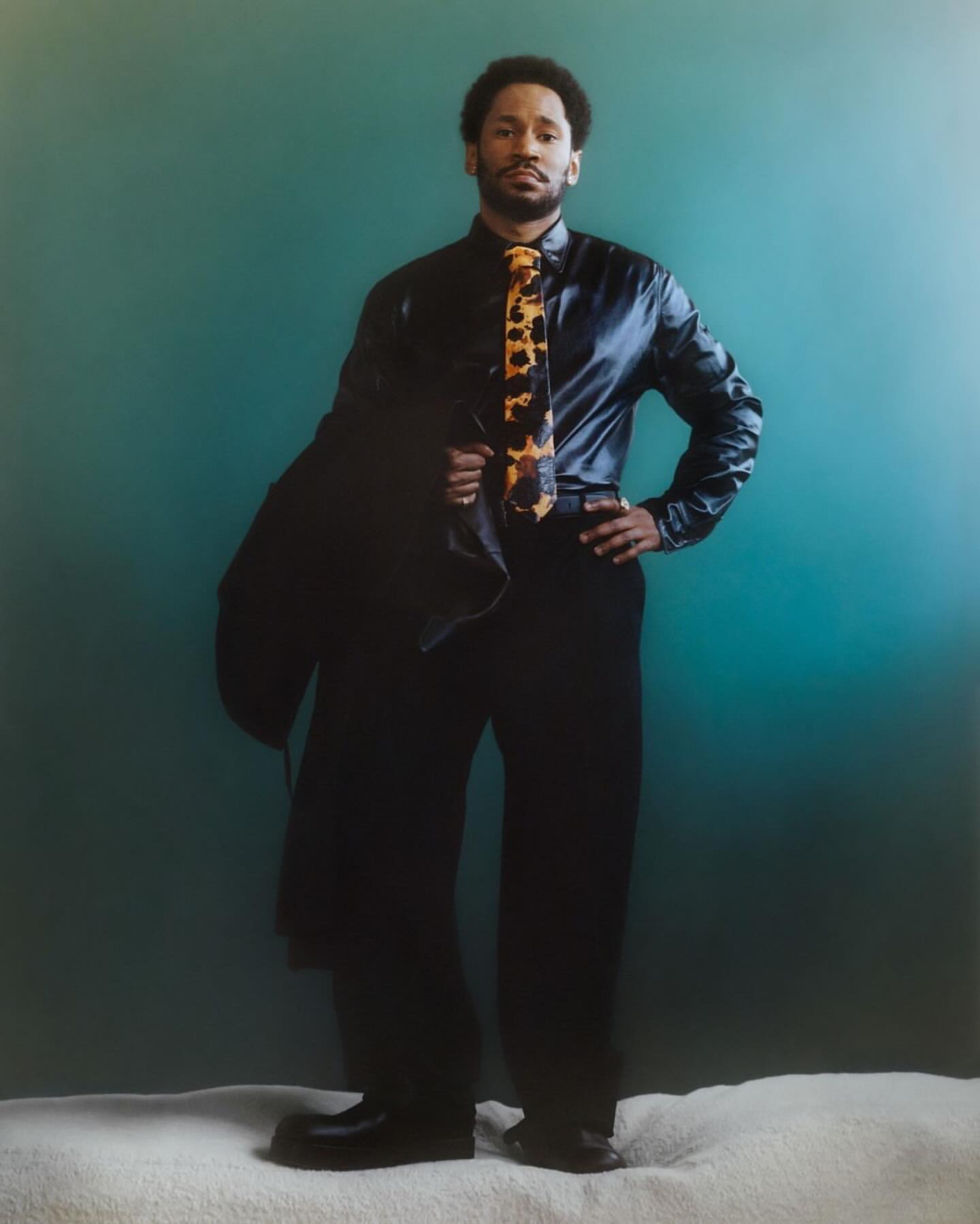 Full body studio shot of an adult male in a black shirt and black pants. He is wearing a patterned tie and holding a jacket.