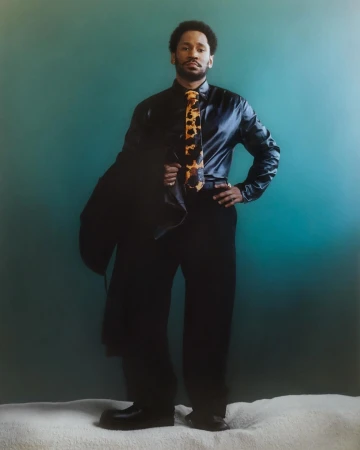 Full body studio shot of an adult male in a black shirt and black pants. He is wearing a patterned tie and holding a jacket.