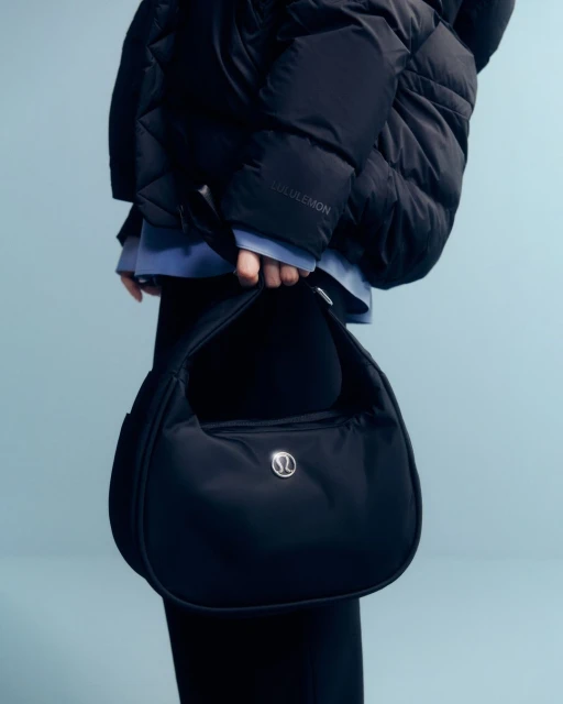 Close-up of a person holding a black Lululemon bag, wearing a puffy black jacket against a light blue backdrop. Fashion shot.