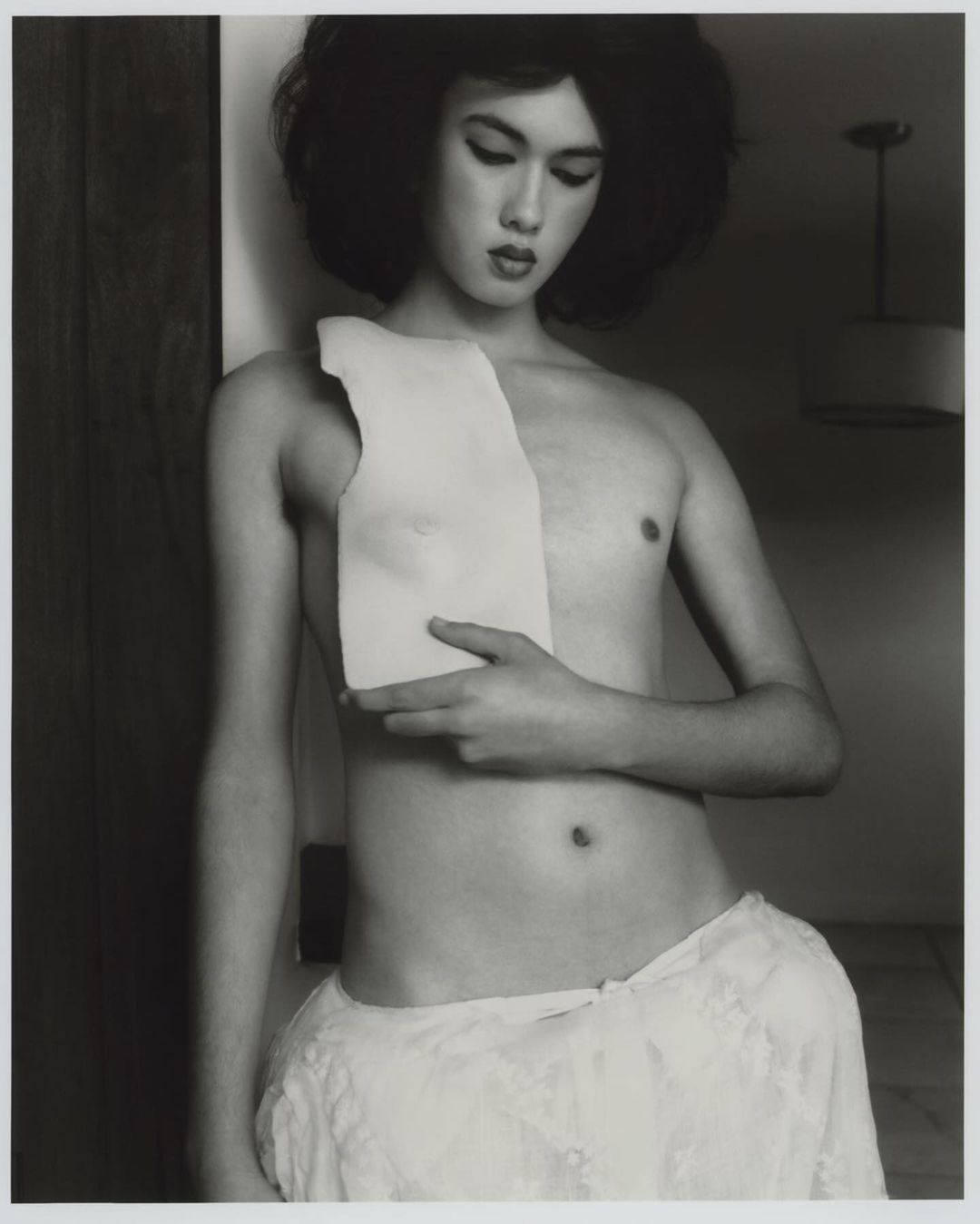 Black and white portrait of a bare-chested person standing with a piece of white material held to their chest in an indoor setting.