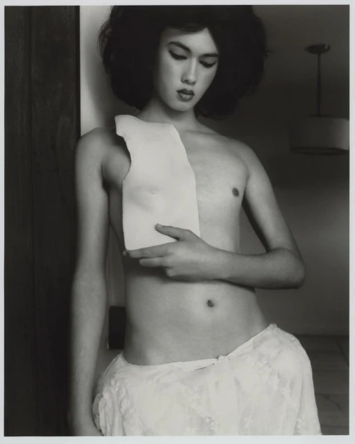 Black and white portrait of a bare-chested person standing with a piece of white material held to their chest in an indoor setting.