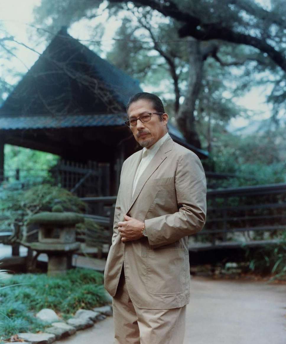 Portrait of a mature adult male in a tan suit and glasses standing outdoors in a garden setting with a wooden structure backdrop.