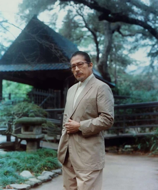 Portrait of a mature adult male in a tan suit and glasses standing outdoors in a garden setting with a wooden structure backdrop.