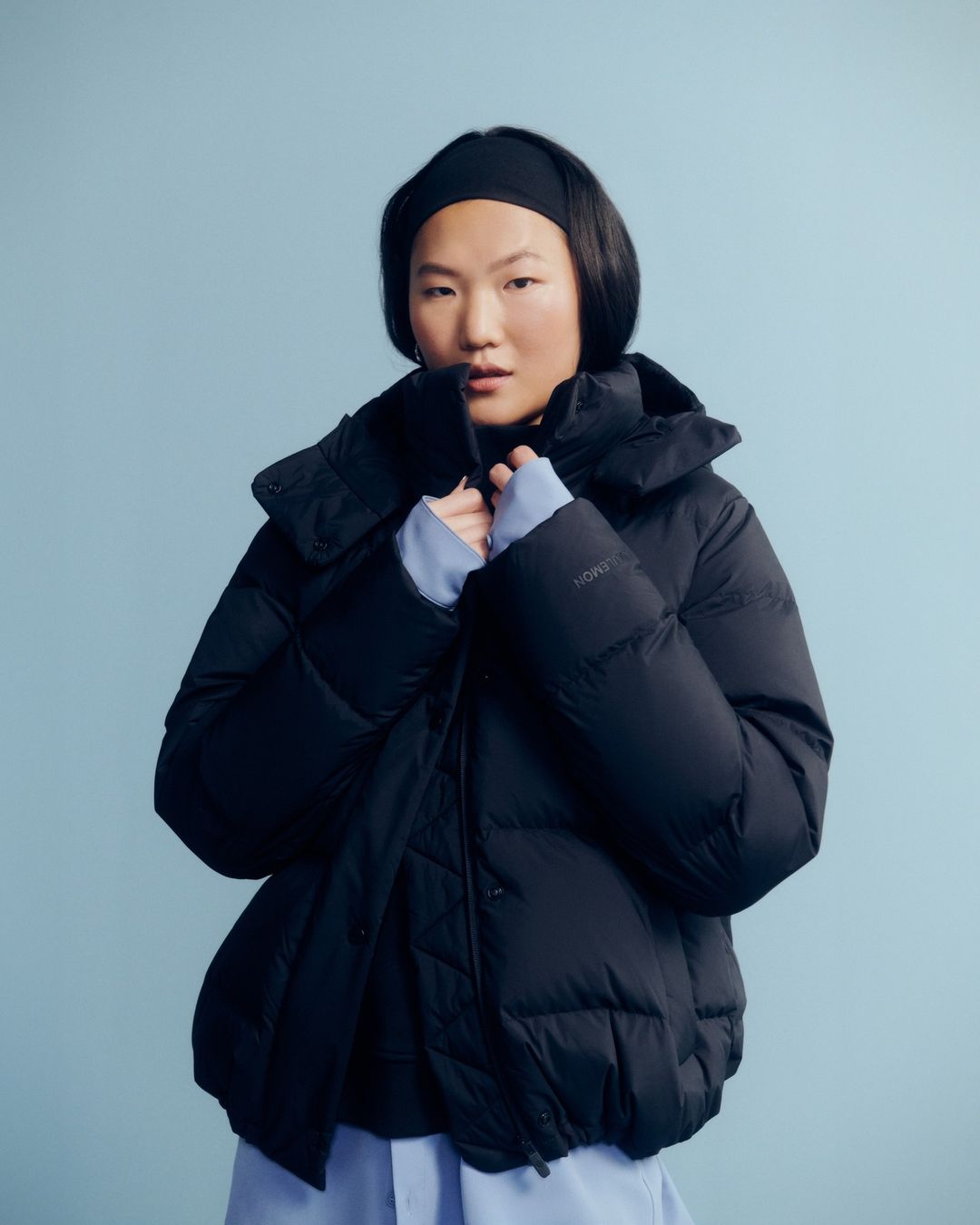Portrait of an Asian woman wearing a black puffer jacket and a matching headband against a solid light blue background in a studio setting.