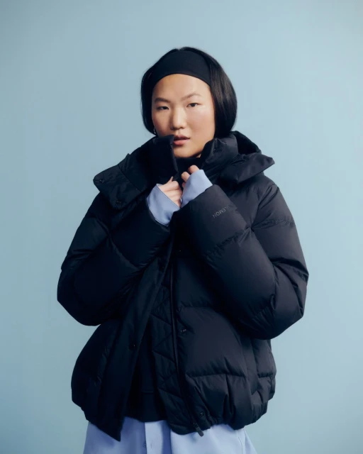 Portrait of an Asian woman wearing a black puffer jacket and a matching headband against a solid light blue background in a studio setting.