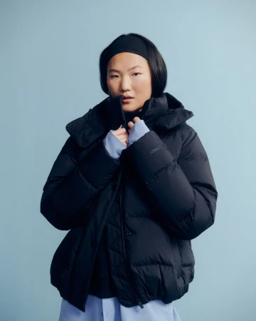 Portrait of an Asian woman wearing a black puffer jacket and a matching headband against a solid light blue background in a studio setting.