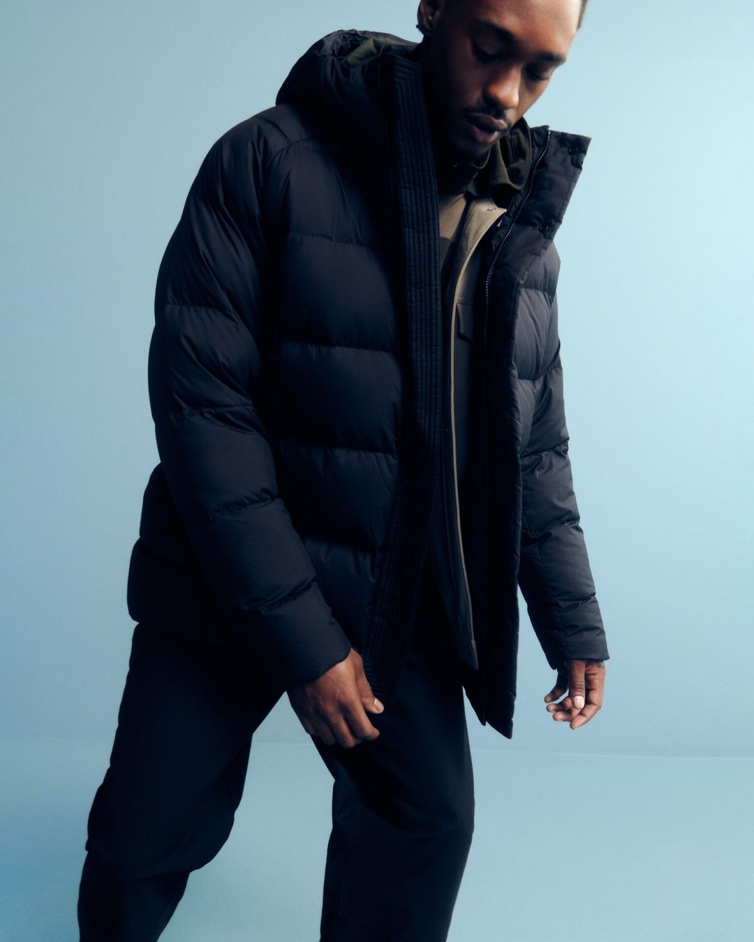 A Black male poses indoors wearing a hooded, black puffer jacket and matching pants against a blue backdrop in a studio shot.