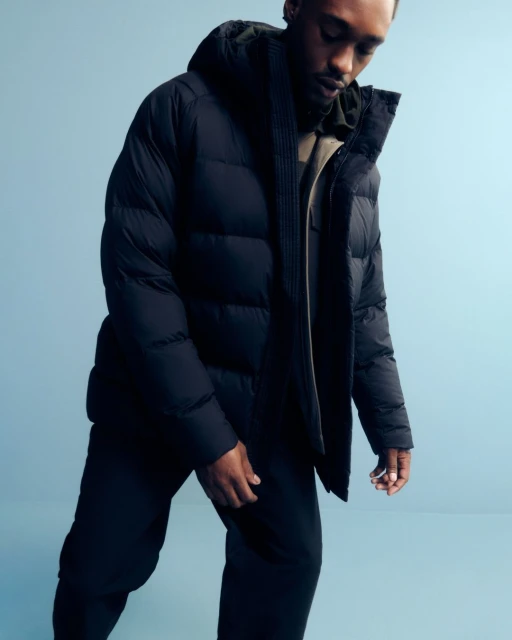 A Black male poses indoors wearing a hooded, black puffer jacket and matching pants against a blue backdrop in a studio shot.