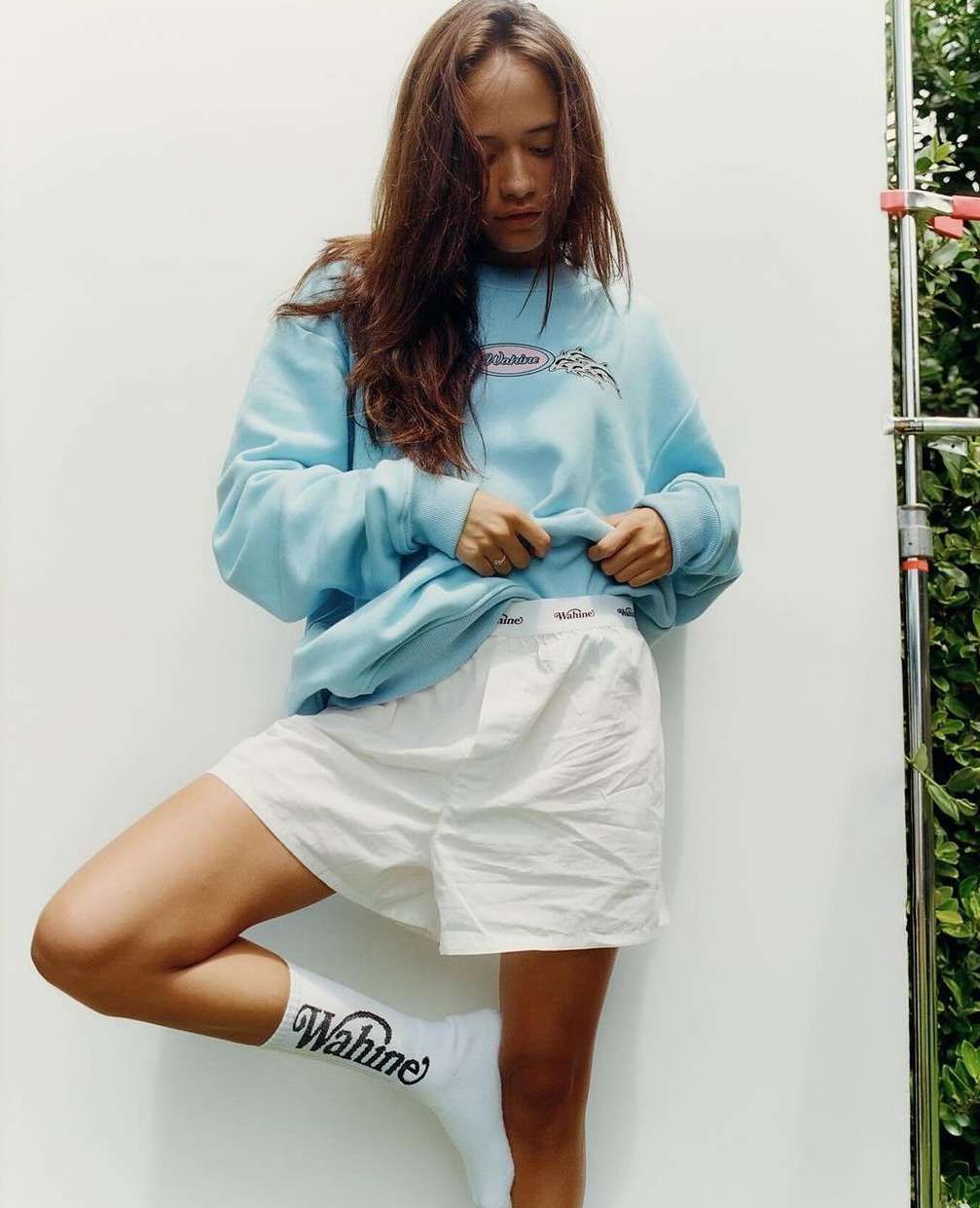 A woman poses in a light blue sweatshirt and white shorts against a white wall with greenery visible in the background, showcasing casual style.