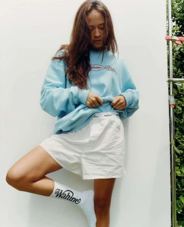 A woman poses in a light blue sweatshirt and white shorts against a white wall with greenery visible in the background, showcasing casual style.