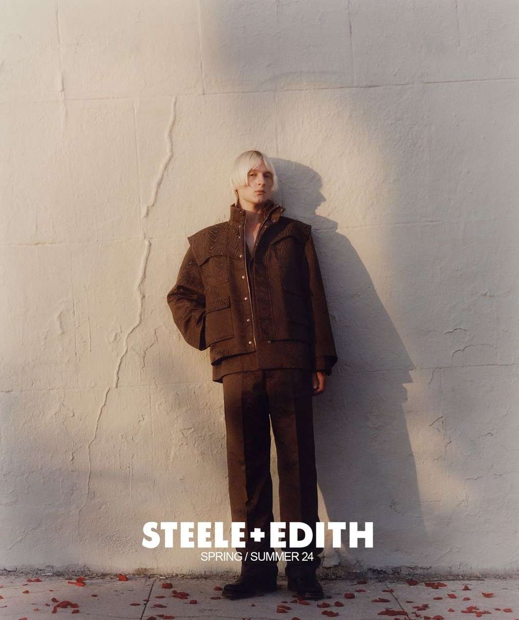 Fashion shot of a young adult with platinum hair, wearing brown corduroy jacket and pants, standing against a textured wall in outdoor lighting.