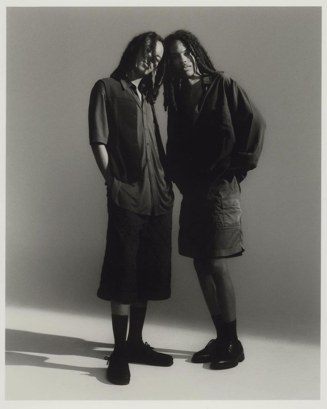 Two black men with dreadlocks wearing dark shirts and shorts standing together against a seamless grey studio backdrop