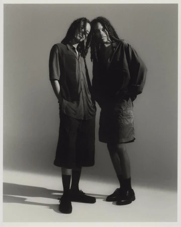 Two black men with dreadlocks wearing dark shirts and shorts standing together against a seamless grey studio backdrop