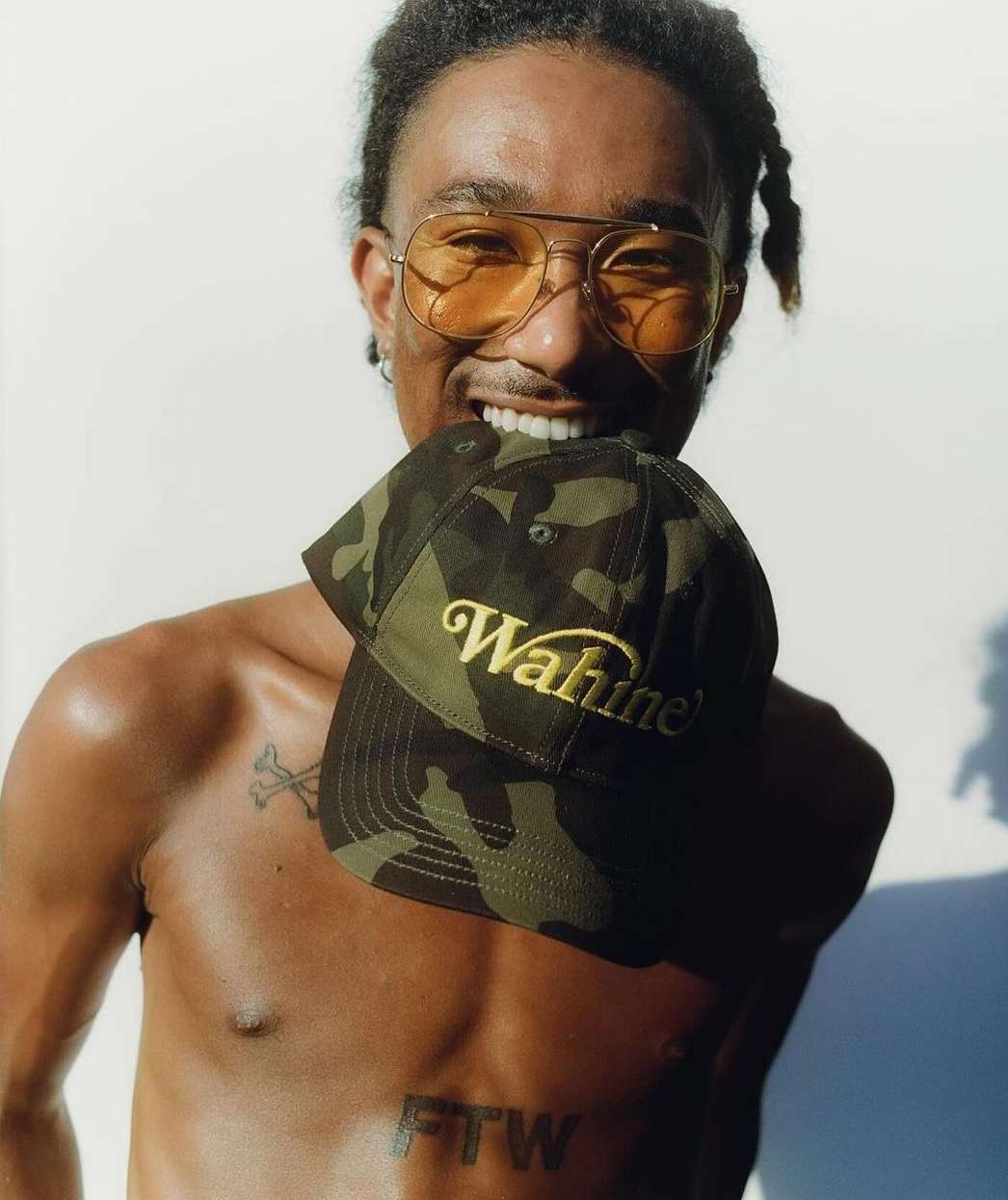 Shirtless African-American man with orange aviator glasses poses with camouflage hat in his mouth against a white background.