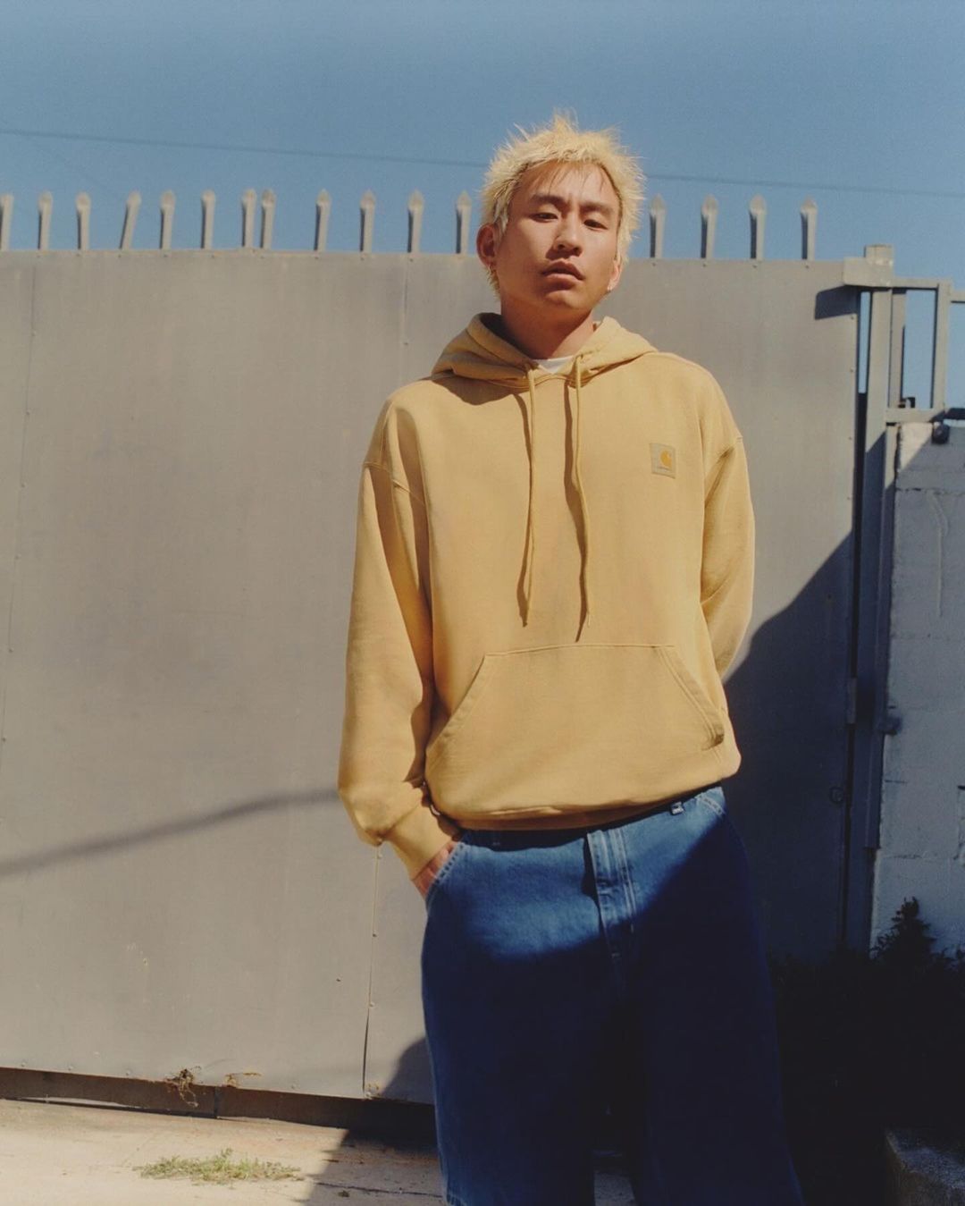Adult male with blonde hair in hooded sweatshirt and jeans poses against grey wall under bright sky in outdoor urban setting.