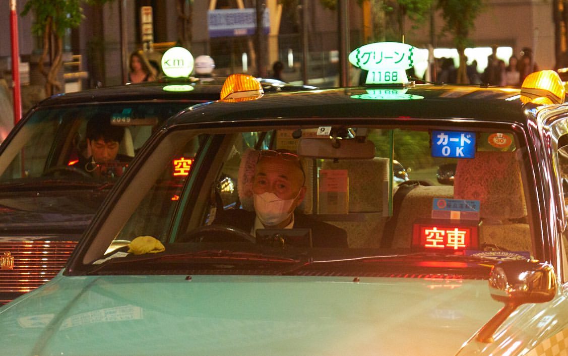Taxi cabs at night in Japan with drivers, one wearing a face mask. City street scene with lights and silhouettes of pedestrians in background.