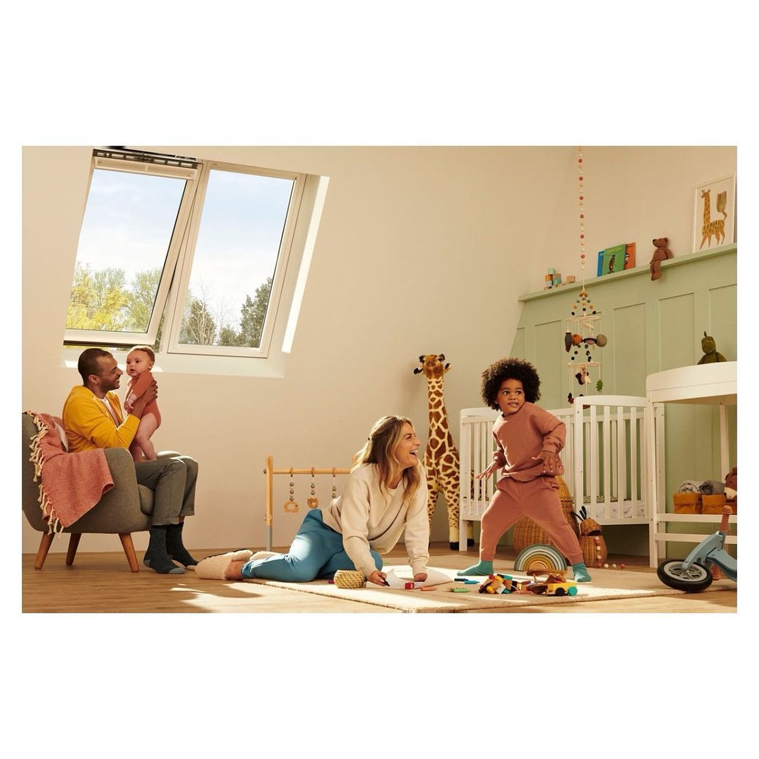 Family plays in a sun-lit attic room: a parent sits with a baby, another plays with a child near a crib under a bright skylight.