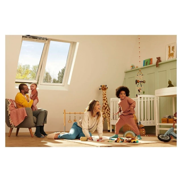 Family plays in a sun-lit attic room: a parent sits with a baby, another plays with a child near a crib under a bright skylight.