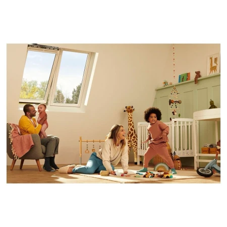Family plays in a sun-lit attic room: a parent sits with a baby, another plays with a child near a crib under a bright skylight.