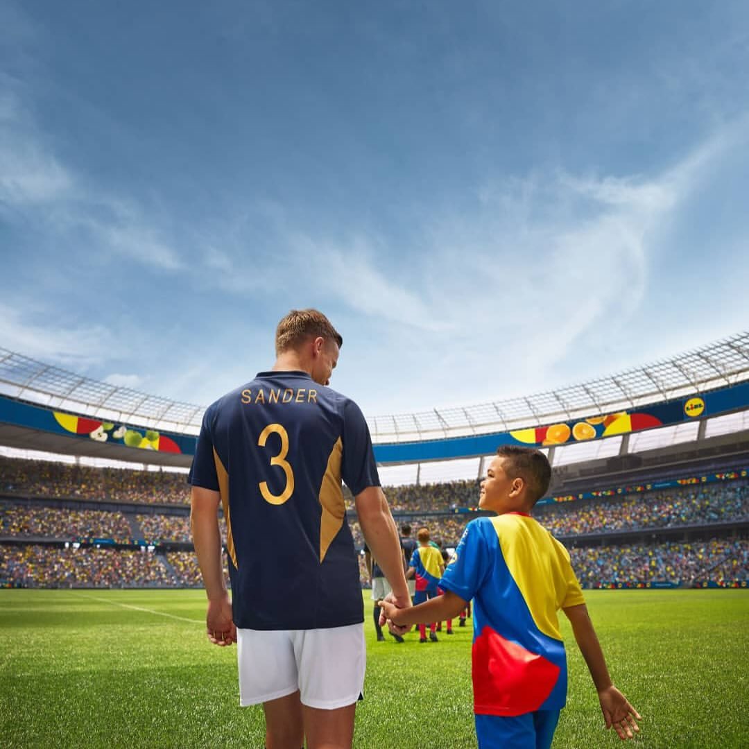 Adult male soccer player in uniform holding hands with a child in a soccer stadium on a sunny day; stadium seating filled with fans.