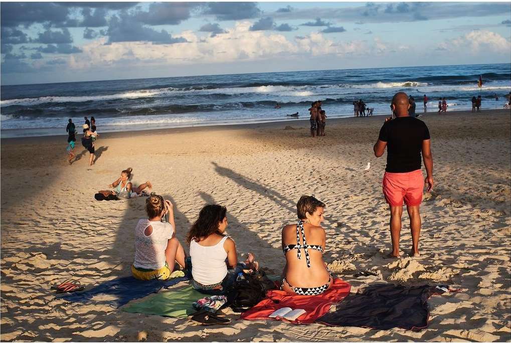 People relaxing and walking on a sandy beach with gentle waves, under a sky with soft clouds during daylight hours.