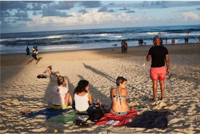 People relaxing and walking on a sandy beach with gentle waves, under a sky with soft clouds during daylight hours. 