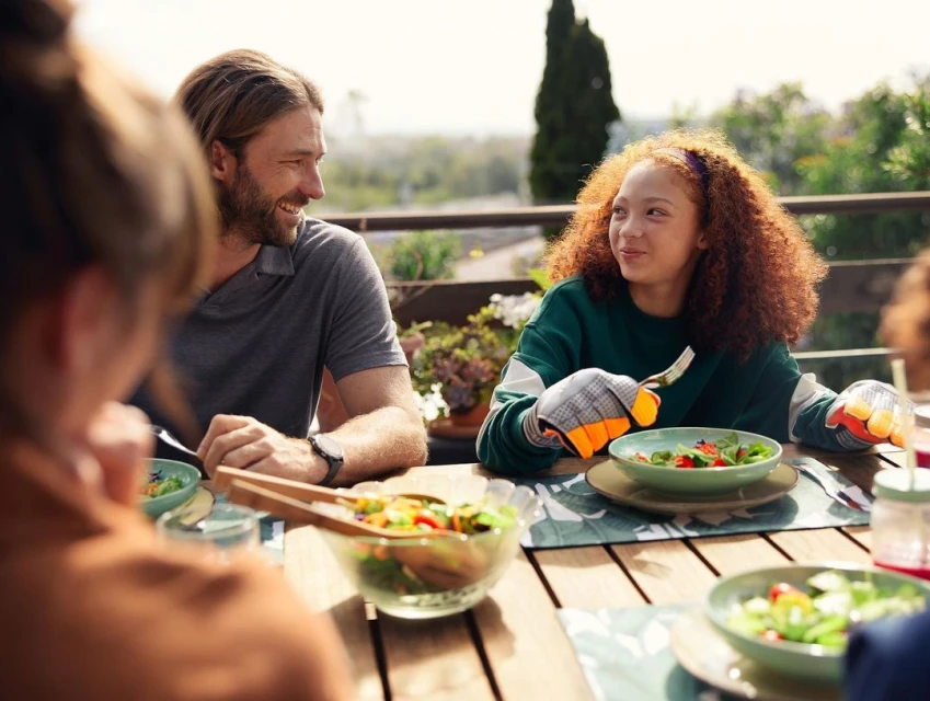 Family shares a salad at an outdoor table on a sunny day. One woman wears gardening gloves and smiles across the table at a man.