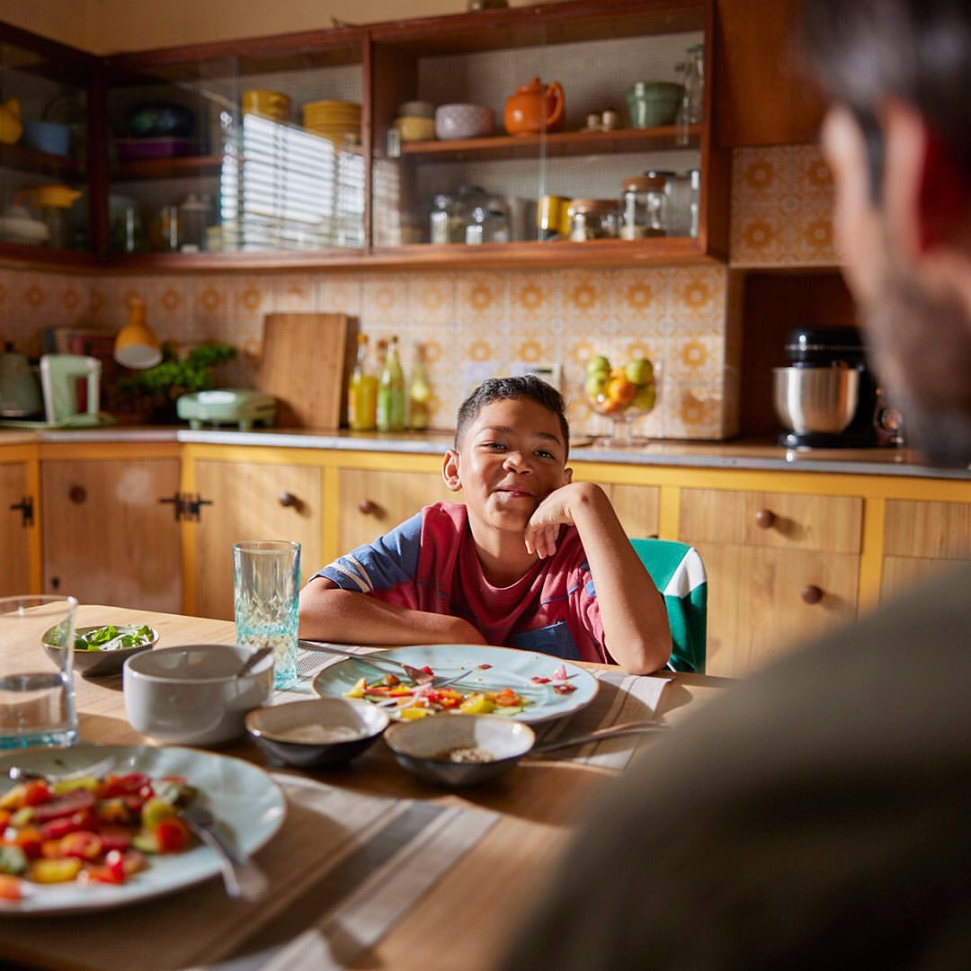A smiling young boy with dark hair and a striped shirt sits at a dining table across from an adult with a meal in a home kitchen.