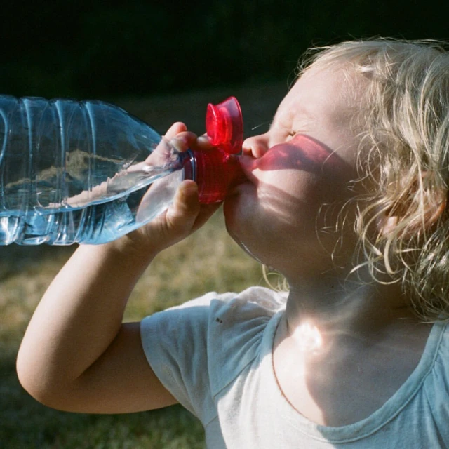 A child drinking from a large clear plastic water bottle with a red lid outdoors, captured in a close-up shot with natural lighting.