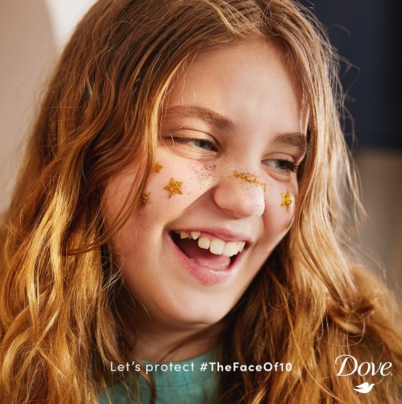 Close-up shot of a smiling pre-teen girl with gold star face decorations and long red hair, against a neutral background.