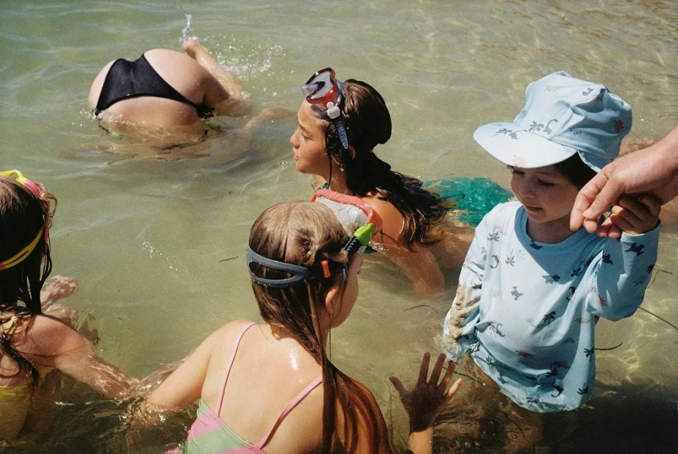 Group of children playing in shallow ocean water on a sunny day, wearing swimsuits, goggles, and a sun hat, enjoying summer vacation.