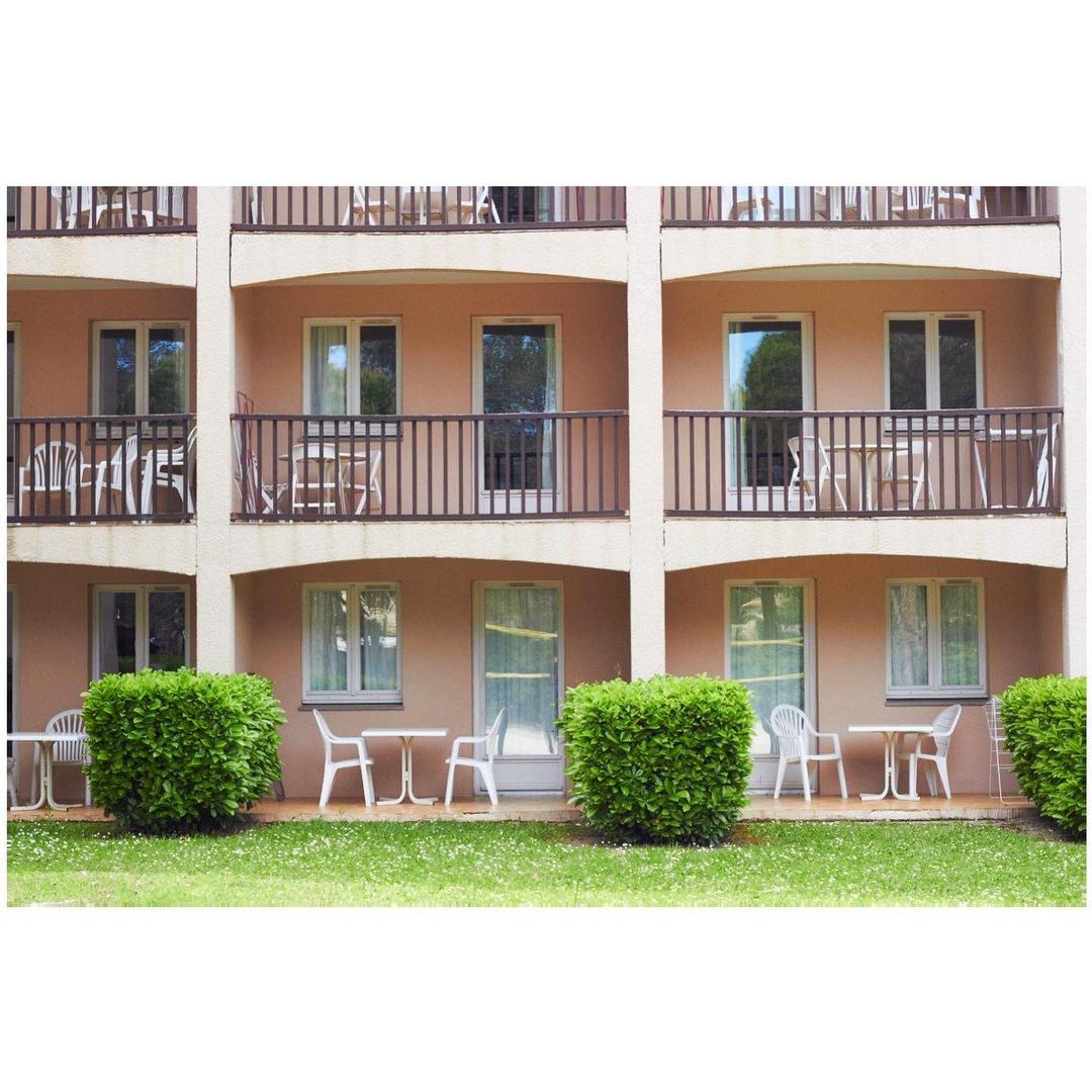 Exterior of a multi-story hotel building with balconies, tables and chairs, and manicured green bushes in the foreground.
